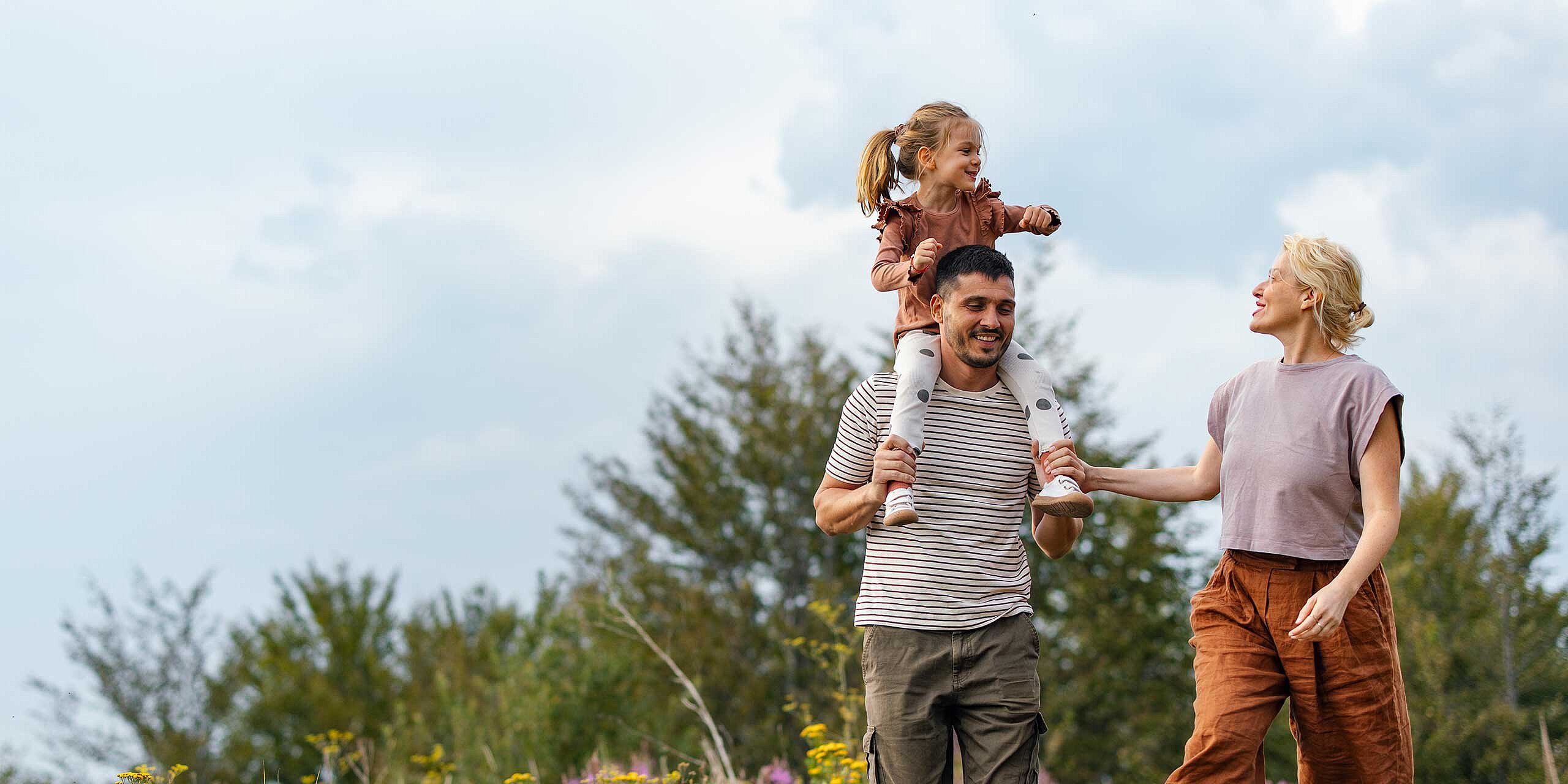 Familie genießt einen Spaziergang im Grünen, das Kind sitzt lachend auf den Schultern des Vaters, während die Mutter daneben geht.