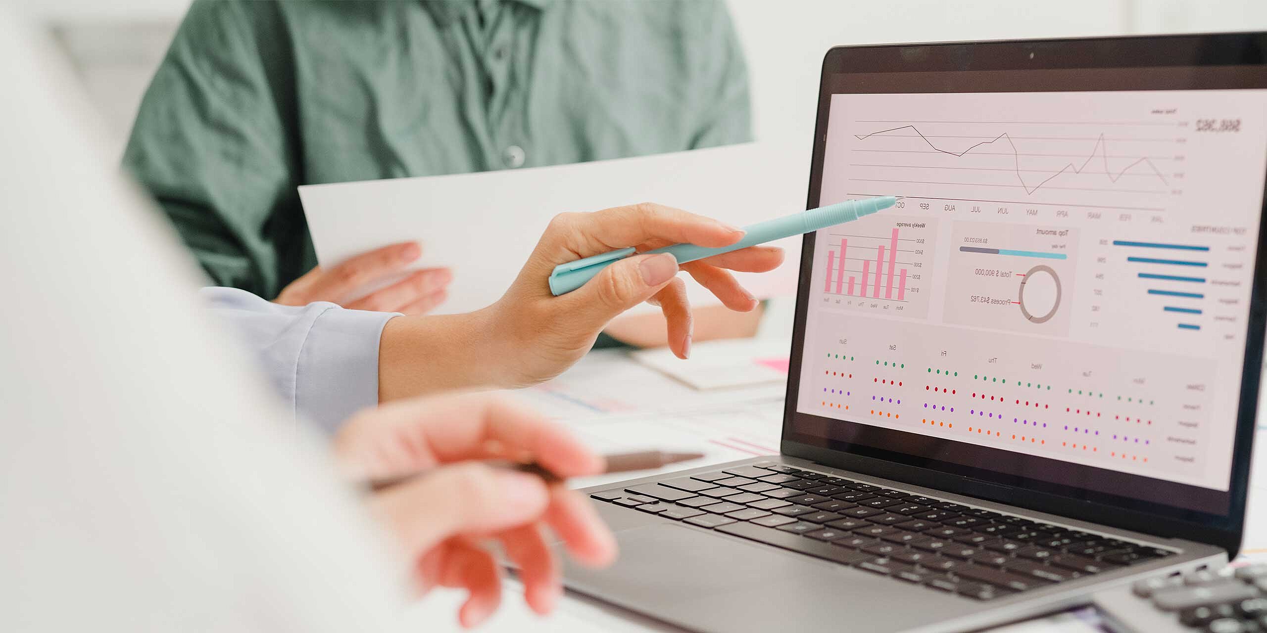 Eine Person zeigt während einer Besprechung Auswertungen und Diagramme auf einem Laptop, während gemeinsam Geschäftszahlen und Kennzahlen analysiert werden.