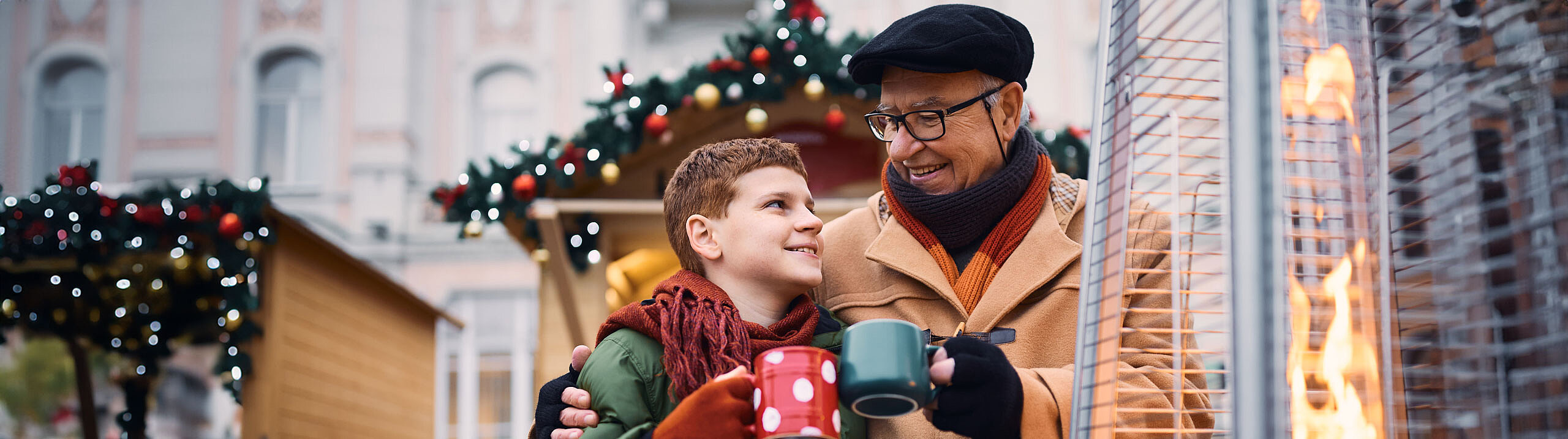 Ein warm gekleideter Mann und ein Kind lächeln, während sie neben einem Außenheizer Becher halten. Sie stehen vor einem festlichen Hintergrund mit dekorierten Holzständen und Weihnachtsgirlanden.