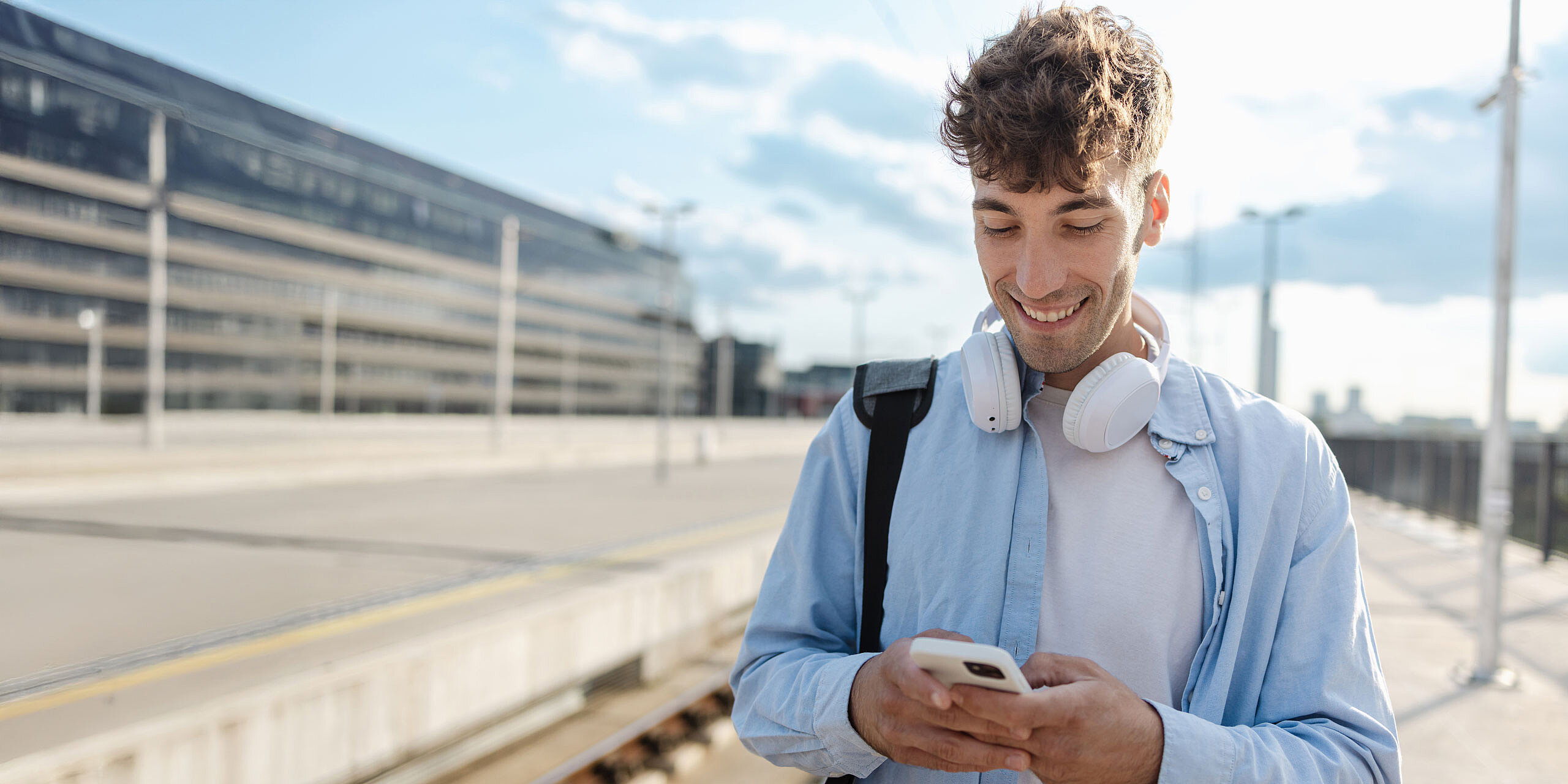 Junger Mann mit Kopfhörern am Bahnhof schaut lächelnd aufs Handy