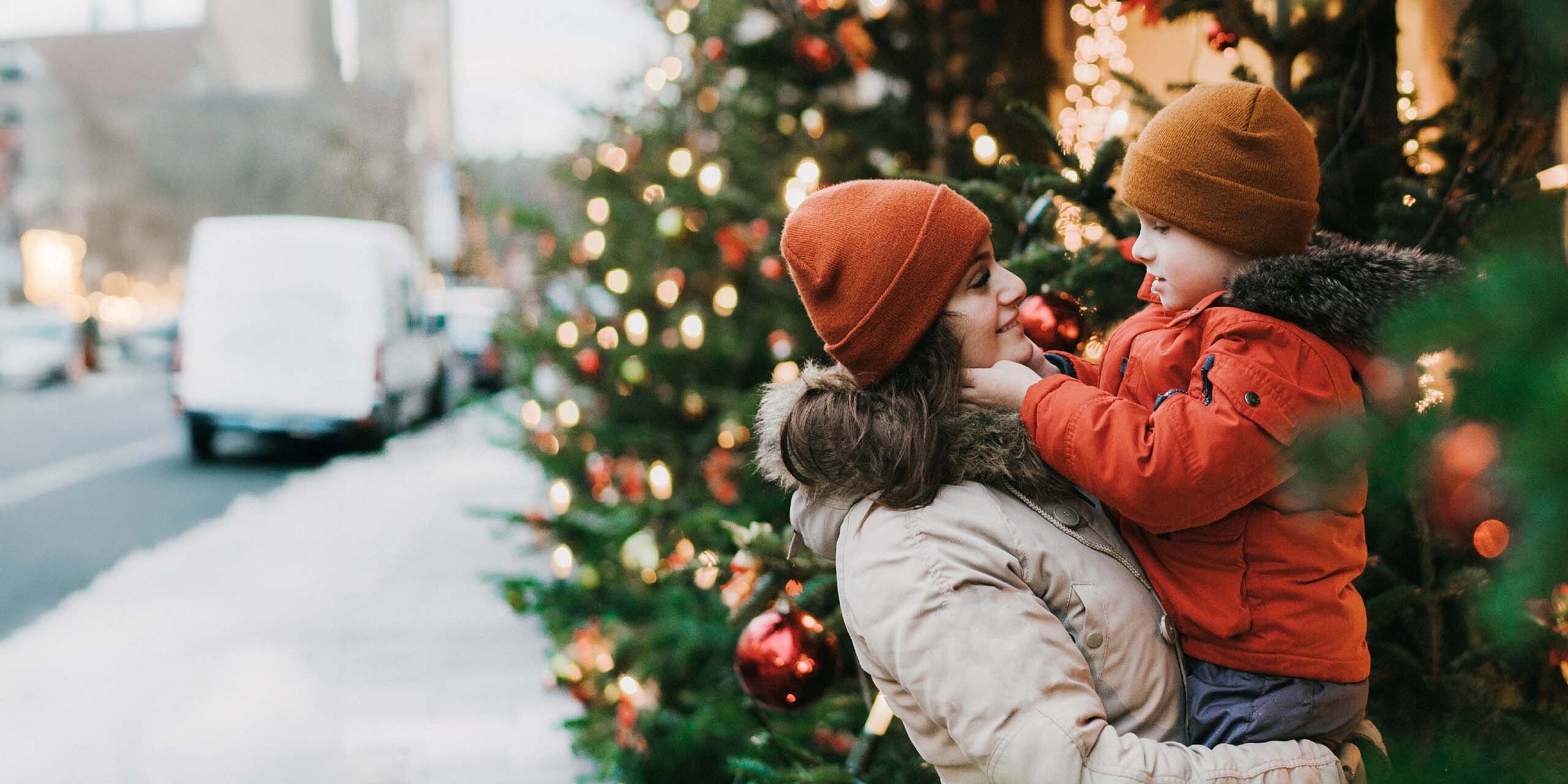 Mutter hält ihr Kind vor weihnachtlich geschmückten Tannenbäumen.