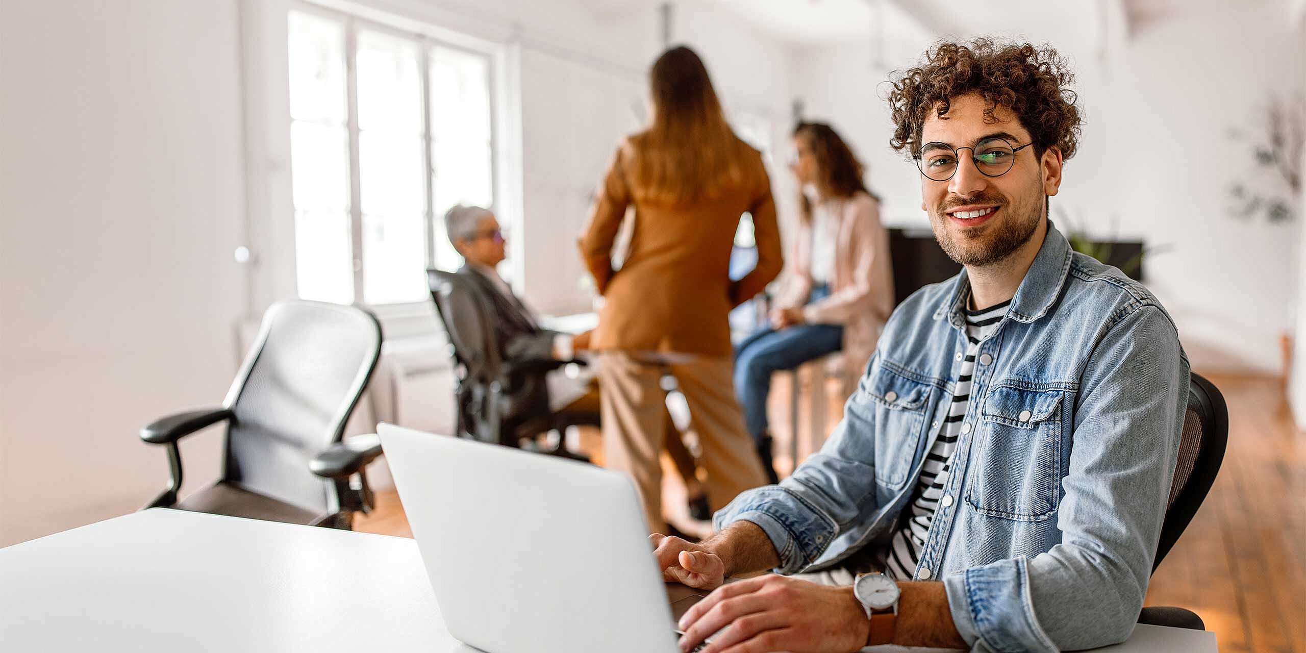 Junger Mann sitzt am Schreibtisch mit Laptop, während ein Team im Hintergrund zusammenarbeitet.