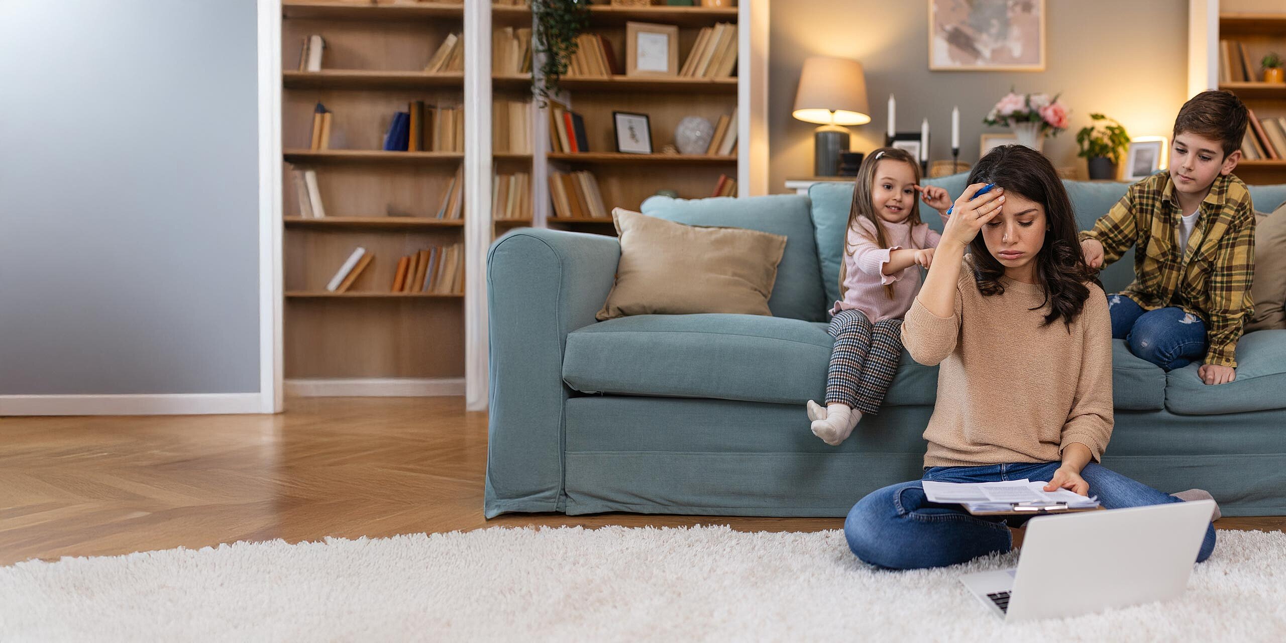 Gestresste Mutter sitzt mit Laptop und Papieren, während Kinder im Hintergrund spielen.