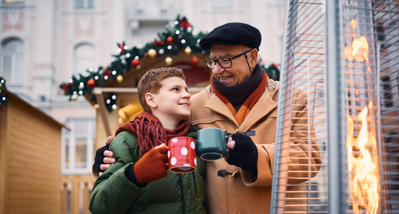 Ein warm gekleideter Mann und ein Kind lächeln, während sie neben einem Außenheizer Becher halten. Sie stehen vor einem festlichen Hintergrund mit dekorierten Holzständen und Weihnachtsgirlanden.