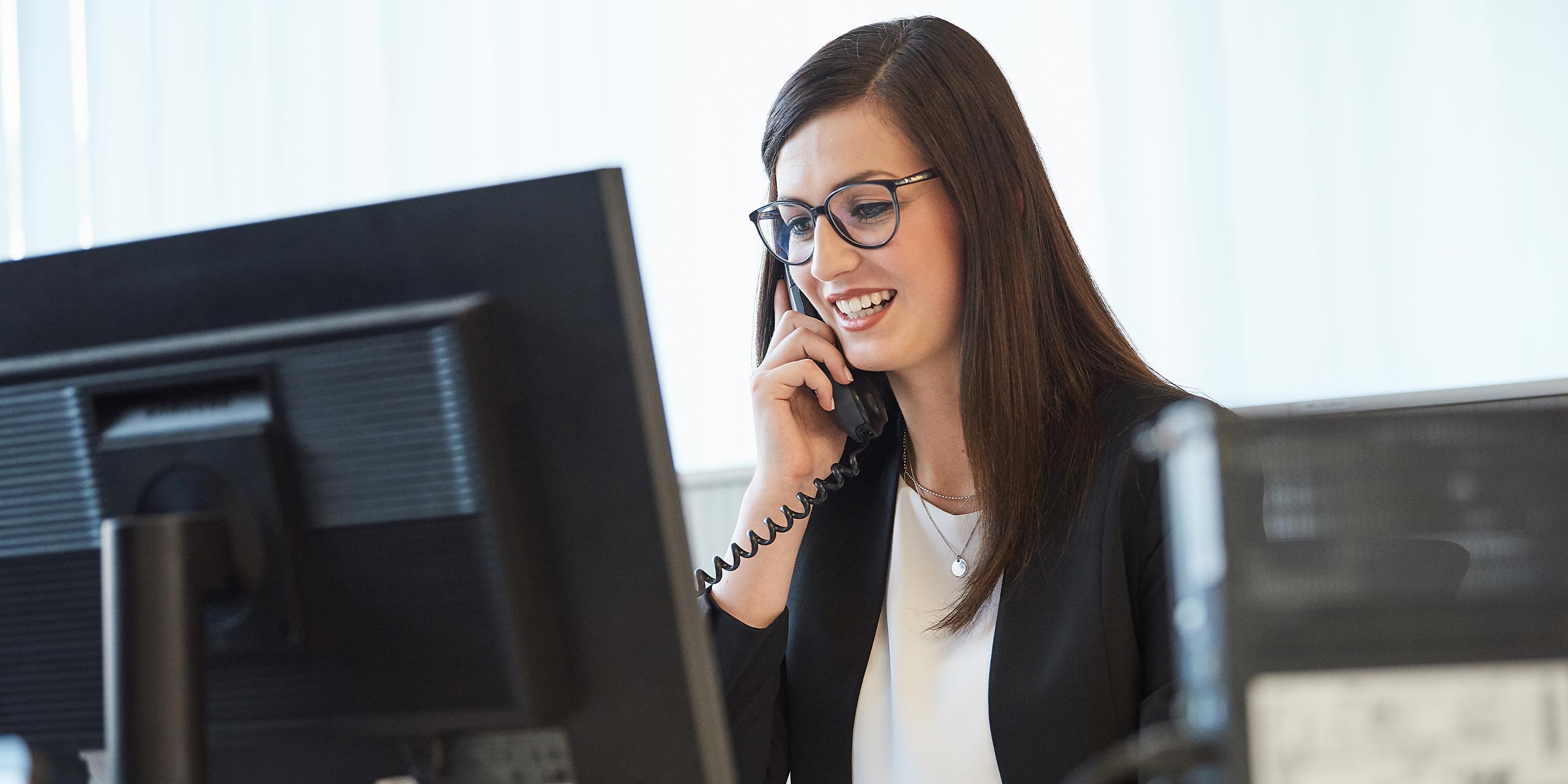 Dunkelhaarige Frau mit Brille sitzt am PC und telefoniert