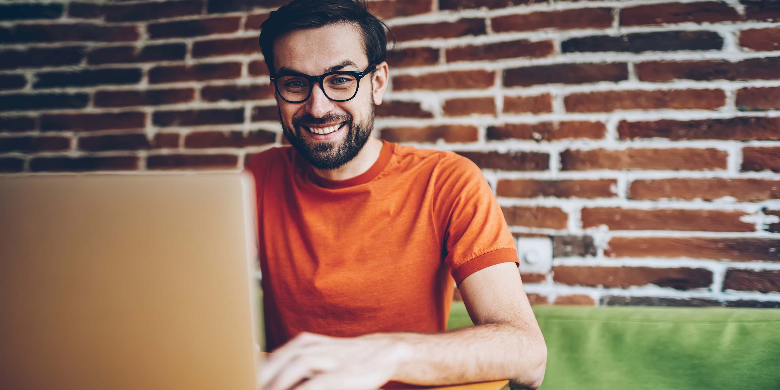 Mann in orangefarbenem T-Shirt schaut lächelnd auf einen Laptop