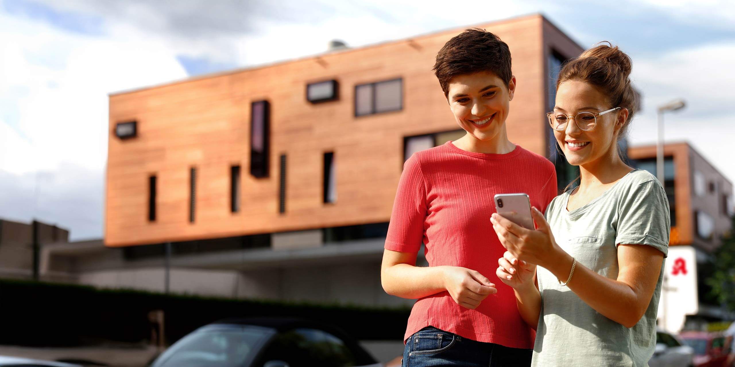 Zwei Frauen stehen draußen in einer modernen städtischen Umgebung. Beide lächeln und schauen auf ein Smartphone, das die Frau rechts hält. Sie sind leger gekleidet und im Hintergrund ist ein modernes Gebäude mit großen Fenstern zu sehen.