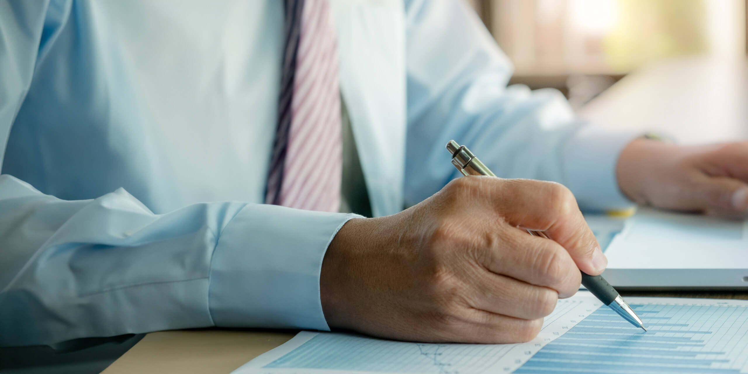 Eine Person in hellblauem Hemd und gestreifter Krawatte sitzt an einem Schreibtisch, hält einen Stift in der Hand und betrachtet ein Finanzdiagramm mit Grafiken. Der Fokus liegt auf der Hand und dem Diagramm, was den Eindruck erweckt, als würde die Person Daten analysieren.
