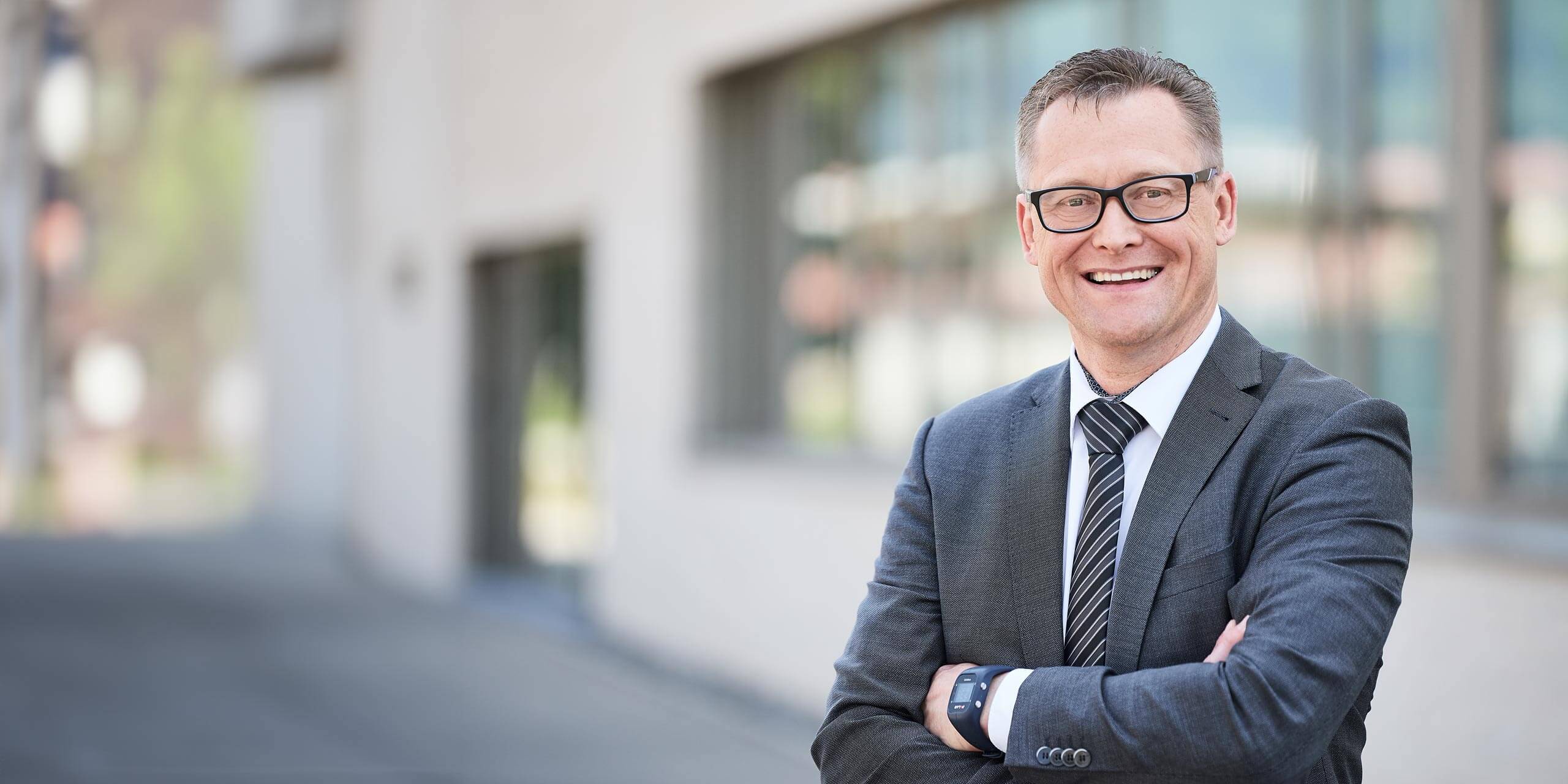 Ein Mann in dunklem Anzug, gestreifter Krawatte und Brille steht lächelnd und mit verschränkten Armen vor einem unscharfen Hintergrund mit modernen Gebäuden im Freien.