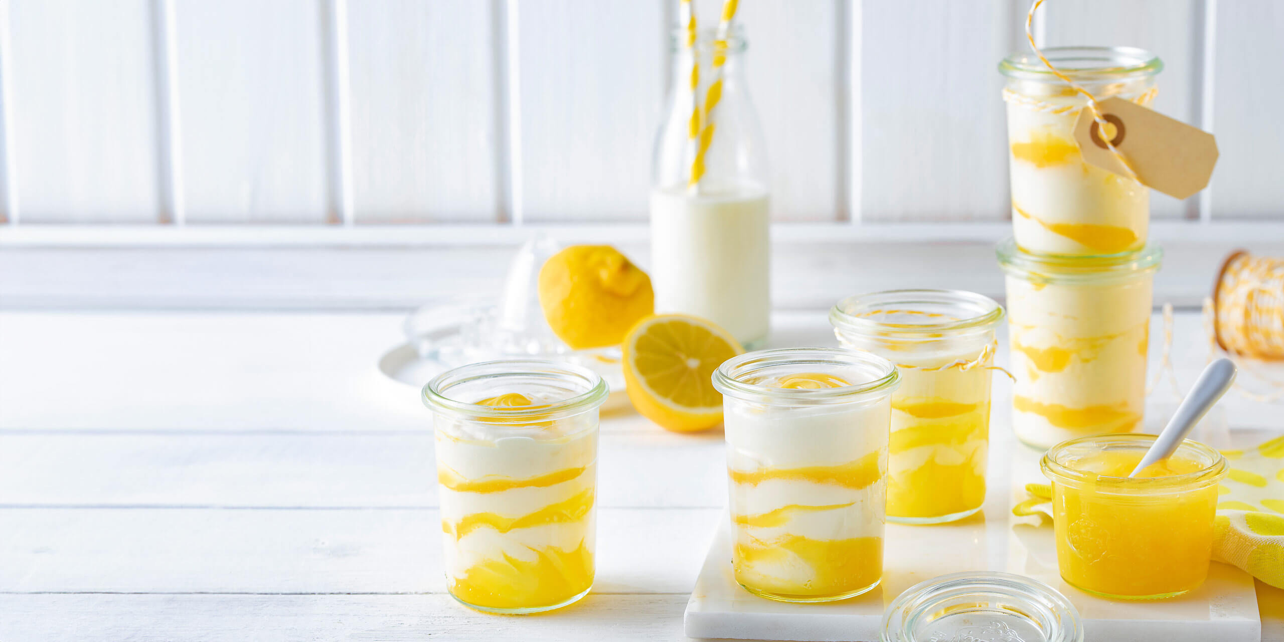 Eine helle und einladende Szene zeigt Gläser mit Zitronen-Joghurt-Parfait, geschichtet mit Zitronenquark und cremigem Joghurt. Eine Flasche Milch mit gelb gestreiftem Strohhalm, frische Zitrone und ein kleines Glas Zitronenquark stehen ebenfalls auf dem weißen Holztisch und vermitteln ein frisches, sommerliches Gefühl.