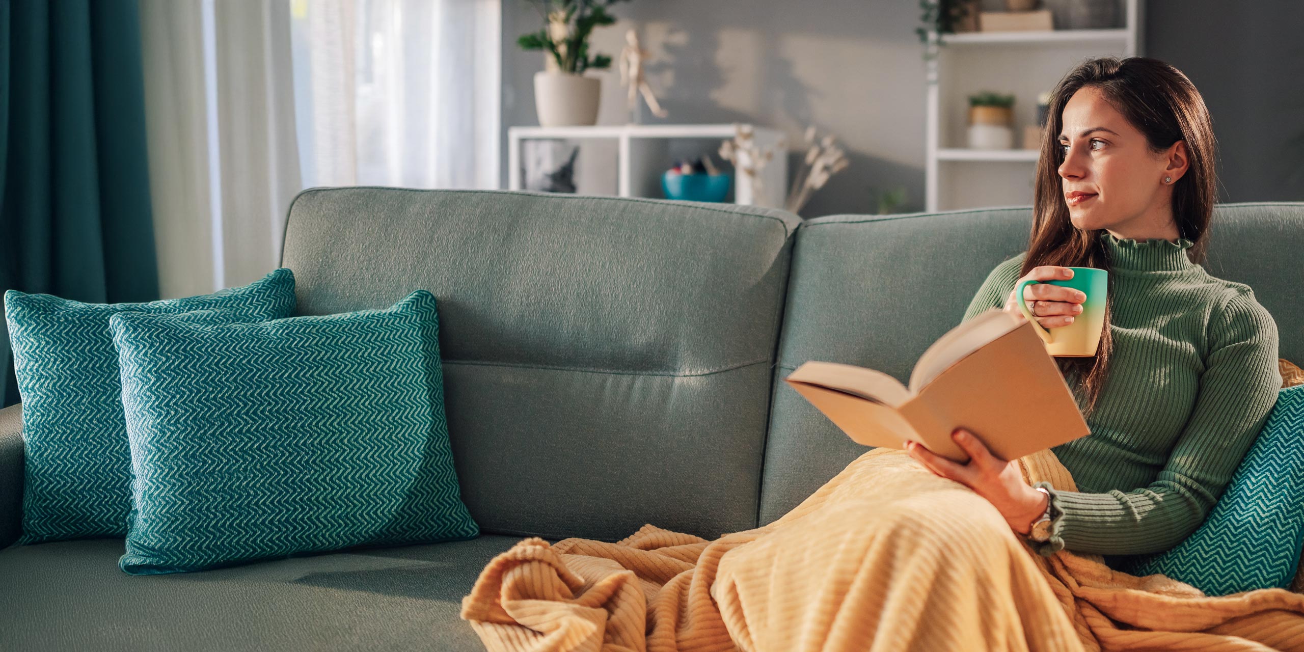 Eine junge Frau mit einer Tasse und einem Buch in der Hand sitzt auf der Couch