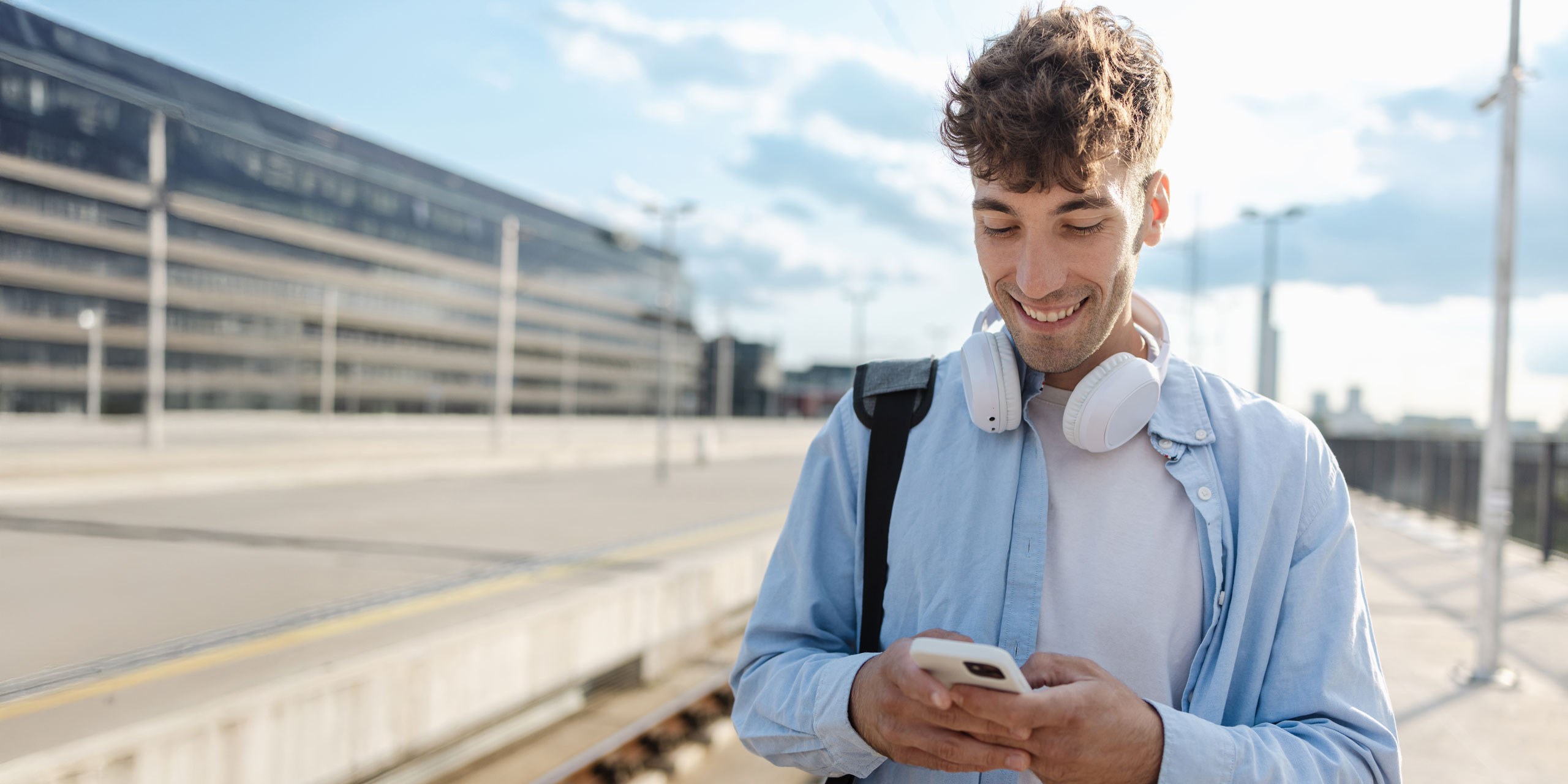 Junger Mann mit Kopfhörern am Bahnhof schaut lächelnd aufs Handy