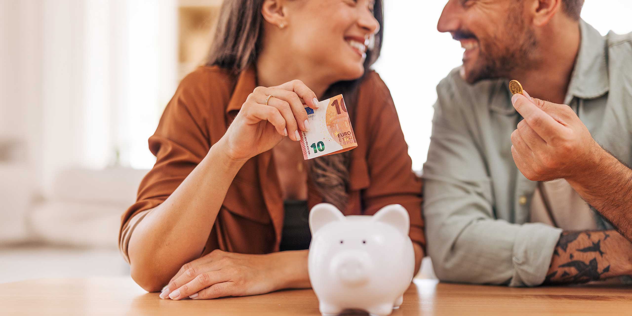 Mann und Frau sparen gemeinsam Geld, vor ihnen steht ein Sparschwein auf dem Tisch.