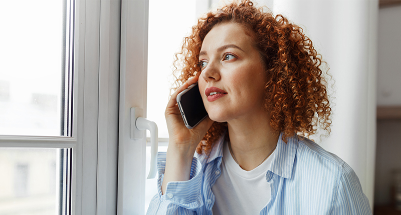 Junge Frau mit Handy am Ohr die aus dem Fenster schaut